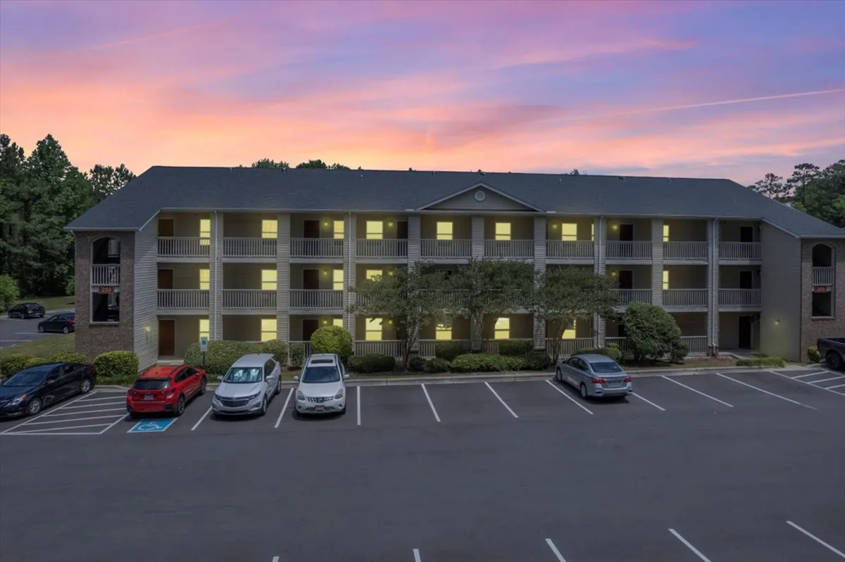 This image captures the apartment building exterior at sunset, with tenants relaxing on balconies, socializing in the parking area, and walking around the property. The well-maintained greenery and open parking lot are also visible, set against a welcoming evening sky.