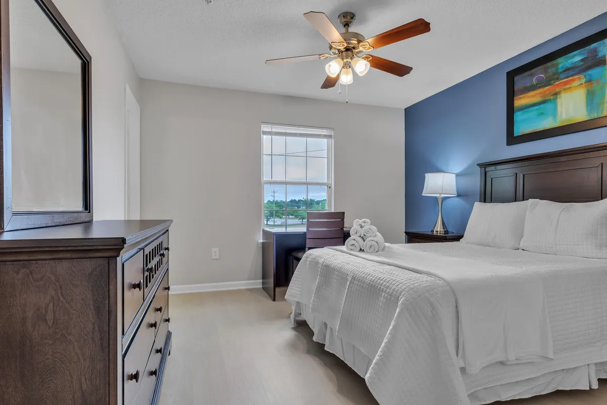 A sunlit bedroom with a bed dressed in crisp white linens, a wooden dresser for storage, and a compact desk with chair positioned near the window. Artwork adds visual interest, and the natural light enhances the sense of peace and space.