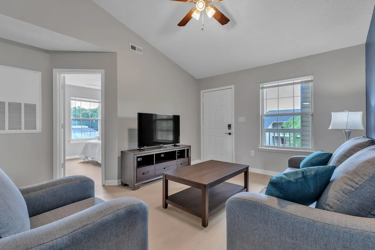 A cozy living space with a plush sofa, coffee table, and a television, positioned near a window that lets in ample natural light. A doorway leads toward a bedroom, giving a glimpse into the apartment's open, connected layout.