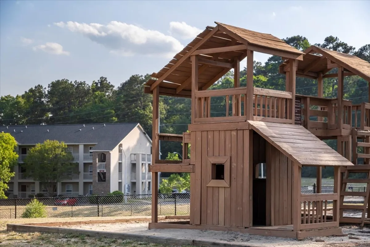 This image captures a lively playground area surrounded by mature trees and a view of the apartment building. Children can be seen playing on the structure, with families gathered around, evoking a joyful atmosphere perfect for group and family activities.