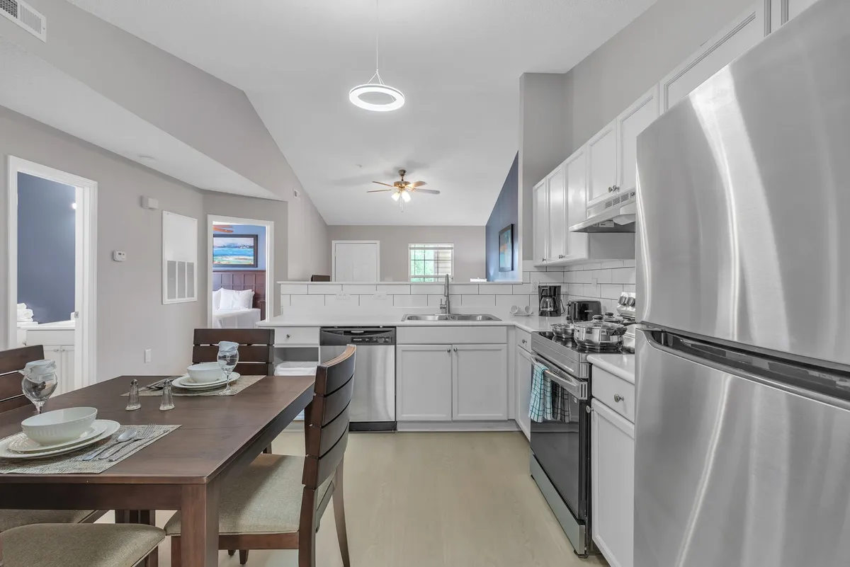 A bright, open dining and kitchen area where the dining table is adjacent to the kitchen appliances, flooded with natural light from the windows. The setup highlights the seamless connection between cooking and gathering spaces, inviting shared meals and togetherness.