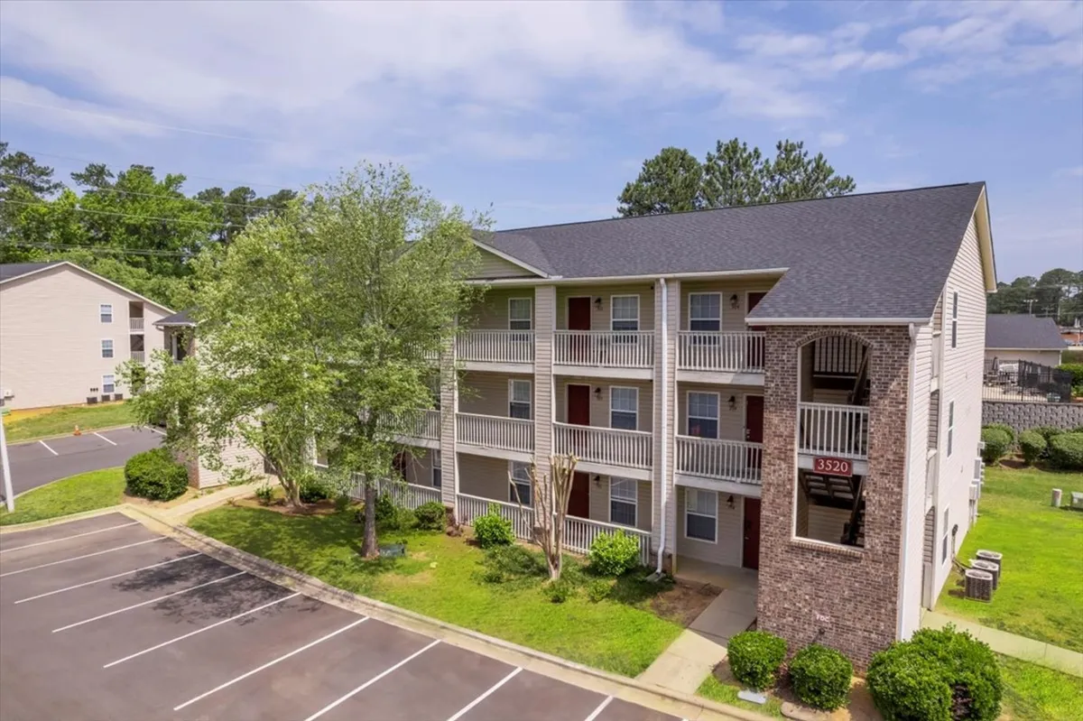 The photo showcases a three-story apartment building surrounded by lush greenery, a paved parking area, and residents enjoying the outdoors. The balconies provide a sense of privacy and relaxation, while the shaded trees create an inviting environment.