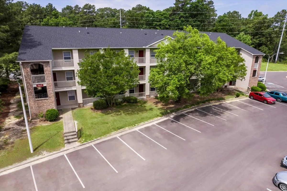 This image shows the exterior of the apartment community with lush green trees, balconies facing the greenery, and the building facade. The parking area is visible, suggesting easy access for residents returning from outings. The scene is bright and inviting, with space for outdoor relaxation or casual gatherings.