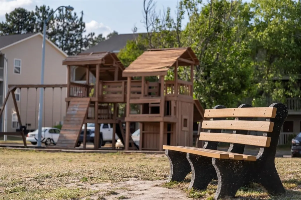 A cheerful exterior scene featuring a wooden playground structure and a park bench, with children playing and families enjoying picnics on the grass. The setting is vibrant and welcoming, encouraging outdoor activities and group adventures.