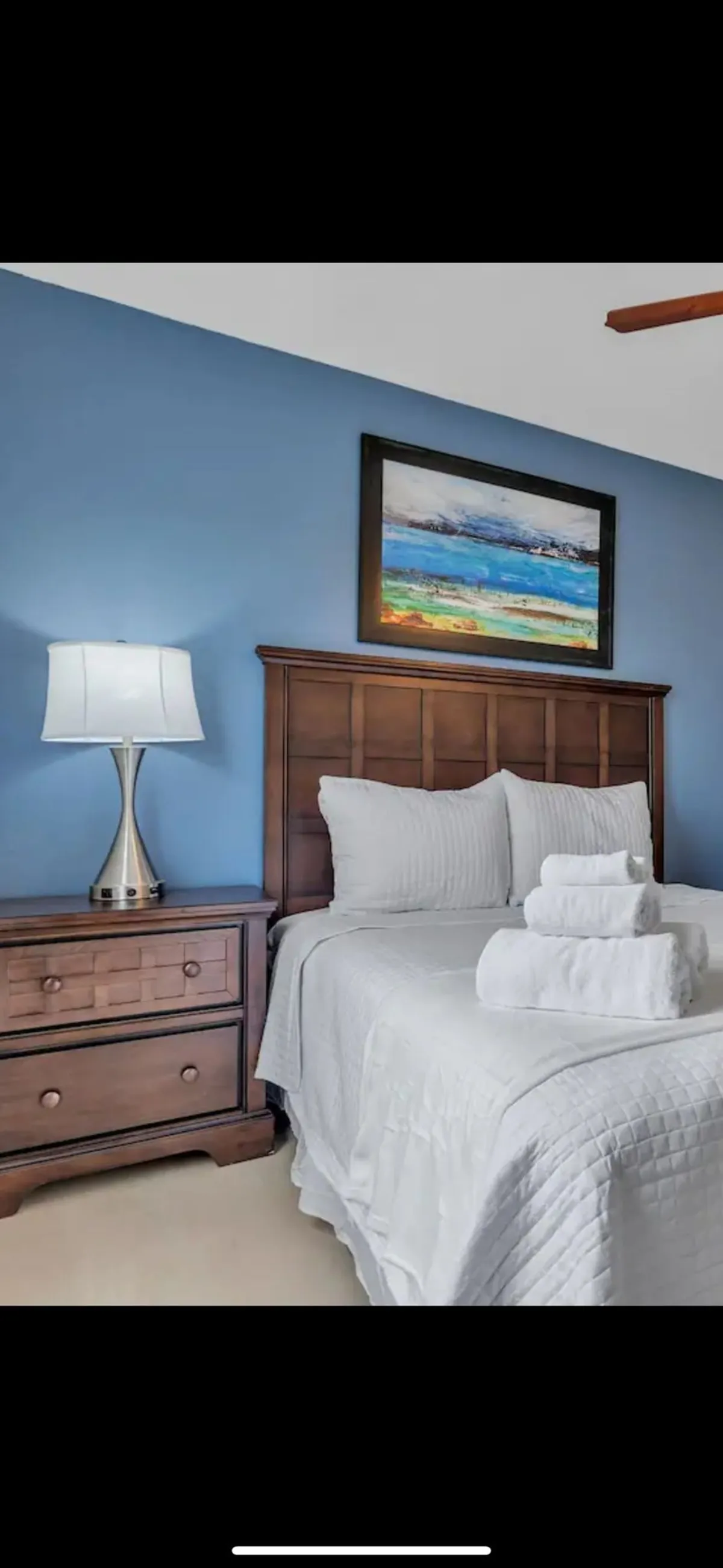 The image showcases a serene bedroom featuring a plush bed with crisp white linens, a warm wooden nightstand topped with a modern lamp, and calming artwork on the wall. The overall atmosphere is tranquil and inviting, ideal for rest and relaxation.